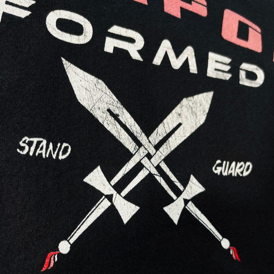 A close up image of a black higher source apparel t shirt with two swords crossed in the center with stand guard in white font.